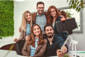 A group of business professionals smiling and posing for a selfie with a smartphone in a modern office setting.