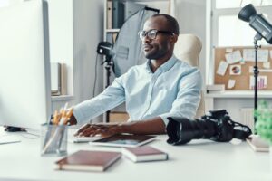 A man with glasses is at a desk, utilizing a camera and computer, immersed in his tasks