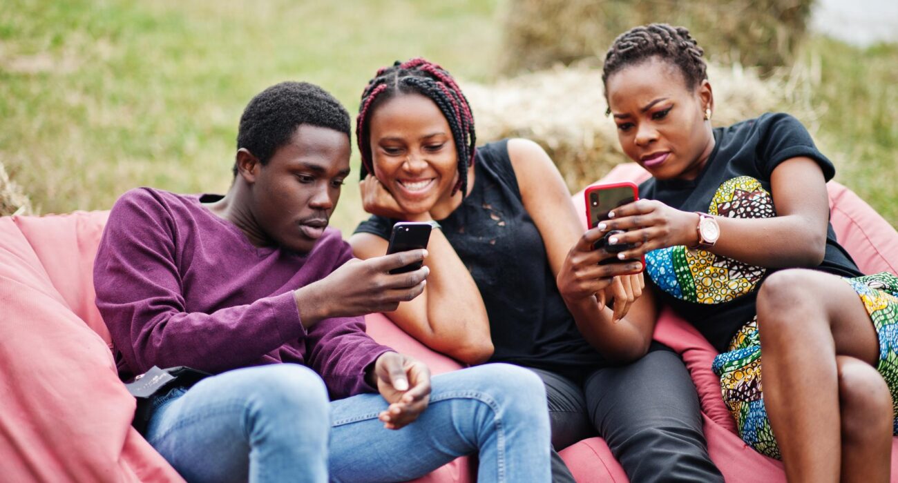 Image of Three african american friends chill, sitting on poufs and using their phones outdoor.